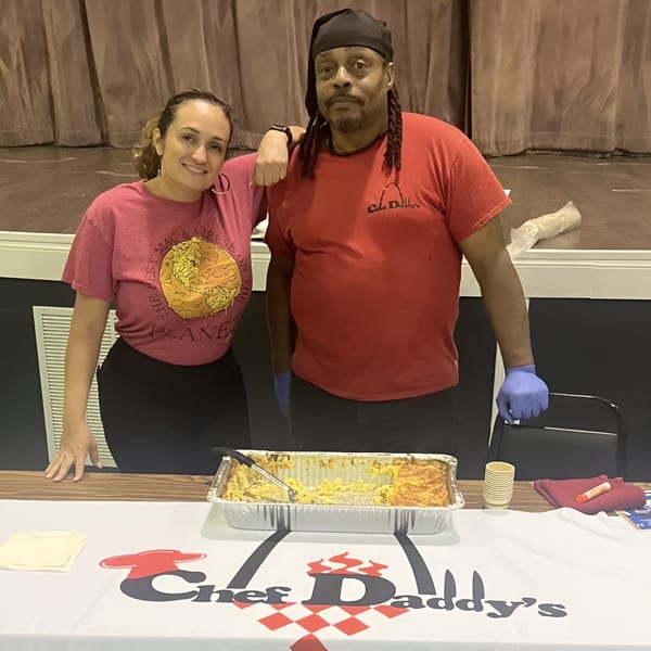 Chef Daddy's owners, a man and woman, stand behind a table with a pan of mac and cheese.