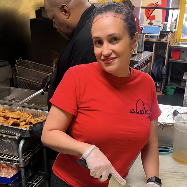 A woman in a red Chef Daddy's shirt smiles while preparing southern fried fish in a kitchen.