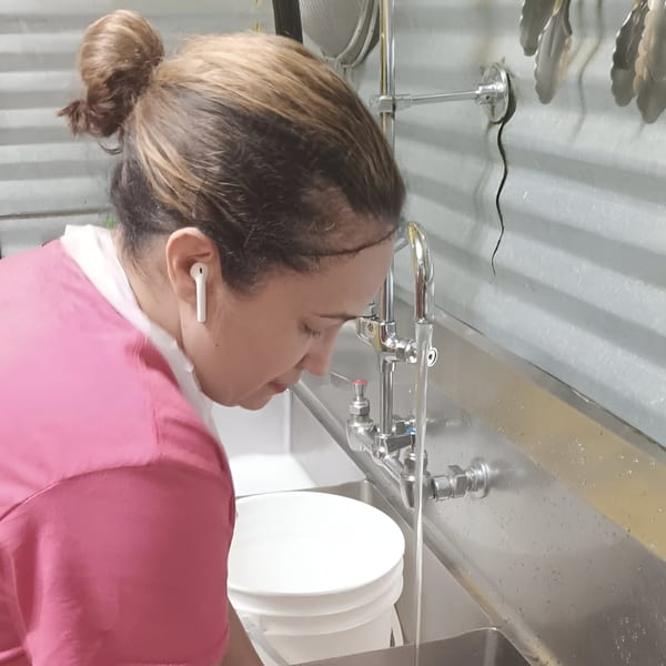 A Chef Daddy's staff member in a pink shirt filling a white bucket at a stainless steel sink.