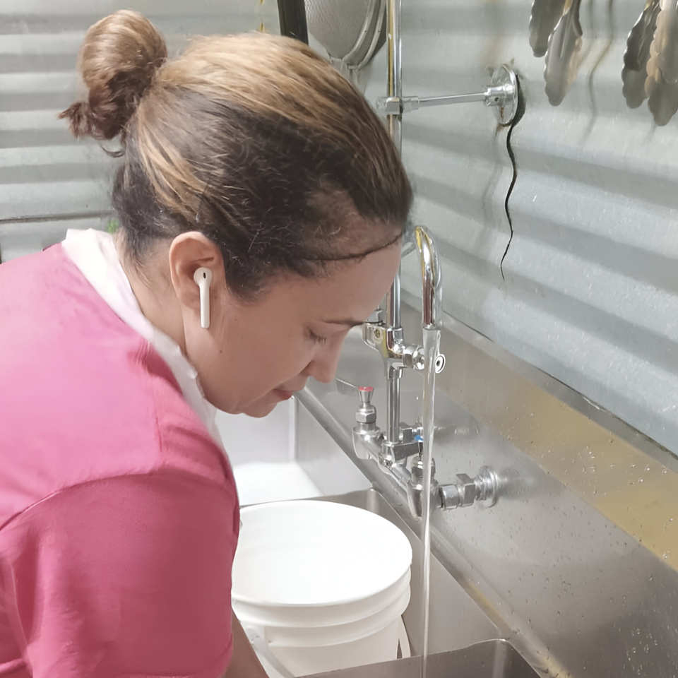 A Chef Daddy's staff member in a pink shirt filling a white bucket at a stainless steel sink.